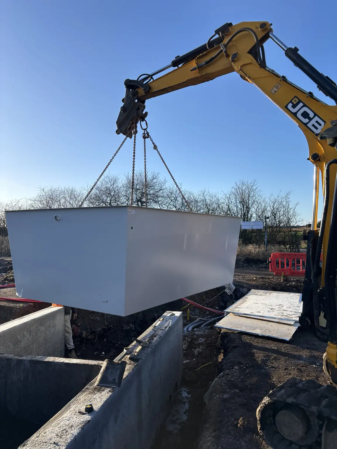 JCB excavator positioning a large electrical enclosure above prepared concrete infrastructure